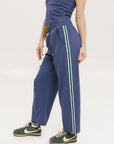 A person standing wearing indigo blue track pants with a drawstring waist and green sneakers.