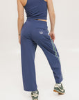 A person standing wearing indigo blue track pants with a drawstring waist and green sneakers.