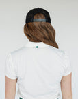 A woman wearing a white golf polo with a collar, a green skirt, and a black hat with text on it.