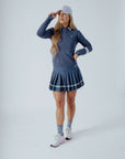 A woman wearing a navy blue 'Dorothy Sweater' with white stripes and an embroidered patch on the chest, paired with a pleated skirt and a white cap.
