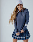 A woman wearing a navy blue 'Dorothy Sweater' with white stripes and an embroidered patch on the chest, paired with a pleated skirt and a white cap.