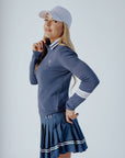 A woman wearing a navy blue 'Dorothy Sweater' with white stripes and an embroidered patch on the chest, paired with a pleated skirt and a white cap.
