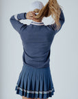 A woman wearing a navy blue 'Dorothy Sweater' with white stripes and an embroidered patch on the chest, paired with a pleated skirt and a white cap.