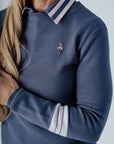 A woman wearing a navy blue 'Dorothy Sweater' with white stripes and an embroidered patch on the chest, paired with a pleated skirt and a white cap.