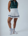 A woman wearing a white pleated golf skirt with a dark green top and white sneakers. Accessories include a tee holder and an embroidered patch on the skirt.