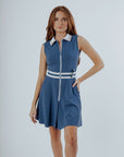 A woman is wearing a sleeveless blue dress with a collar and a front zipper detail. The dress features a contrasting white and green trim and she has a pocket on the hip.