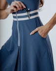 A woman is wearing a sleeveless blue dress with a collar and a front zipper detail. The dress features a contrasting white and green trim and she has a pocket on the hip.