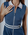 A woman is wearing a sleeveless blue dress with a collar and a front zipper detail. The dress features a contrasting white and green trim and she has a pocket on the hip.