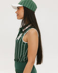 A female model wearing a sleeveless green pinstripe polo top with an applique logo on the chest and a matching green skirt, paired with a green cap.