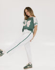 A woman posing with one leg lifted, wearing a green and white t-shirt with the text 'CHASING BALLS' on the back, paired with white pants and green sneakers.