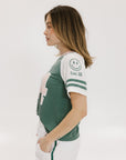 A woman wearing a green and white boxy fit t-shirt with the number 04 on the front and bold lettering on the back, along with striped sleeves.