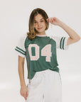 A woman wearing a green and white boxy fit t-shirt with the number 04 on the front and bold lettering on the back, along with striped sleeves.