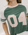 A woman wearing a green and white boxy fit t-shirt with the number 04 on the front and bold lettering on the back, along with striped sleeves.