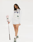 A woman wearing a white sleeveless tank top with 'Fore All' text and a flamingo logo on the chest, paired with a white skirt.