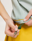 A woman wearing a green striped seersucker polo top with a collar, sleeveless with front yoke and piping accents, paired with a yellow skirt and a matching green cap.