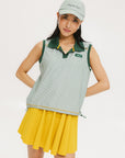 A woman wearing a green striped seersucker polo top with a collar, sleeveless with front yoke and piping accents, paired with a yellow skirt and a matching green cap.