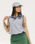 A woman wearing a sleeveless indigo-striped seersucker polo shirt with collar, sleeve yoke, and piping accents. The shirt has elastic toggles at the hem and side seam, and sleeveless appliqué logos.