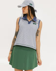 A woman wearing a sleeveless indigo-striped seersucker polo shirt with collar, sleeve yoke, and piping accents. The shirt has elastic toggles at the hem and side seam, and sleeveless appliqué logos.