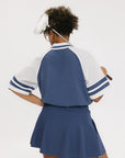 A person wearing a blue and white rugby-inspired polo shirt with Mesh body appliqué logos, a ribbed collar and placket, and elastic toggles at the side seam hem. The shirt has 'FORE ALL' text and a golf club logo on the front.