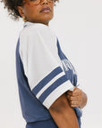 A person wearing a blue and white rugby-inspired polo shirt with Mesh body appliqué logos, a ribbed collar and placket, and elastic toggles at the side seam hem. The shirt has 'FORE ALL' text and a golf club logo on the front.