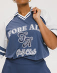 A person wearing a blue and white rugby-inspired polo shirt with Mesh body appliqué logos, a ribbed collar and placket, and elastic toggles at the side seam hem. The shirt has 'FORE ALL' text and a golf club logo on the front.