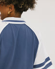 A person wearing a blue and white rugby-inspired polo shirt with Mesh body appliqué logos, a ribbed collar and placket, and elastic toggles at the side seam hem. The shirt has 'FORE ALL' text and a golf club logo on the front.