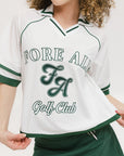 A woman wearing a white rugby-inspired polo shirt with green details and text that reads 'FORE ALL Golf Club'.