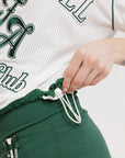 A woman wearing a white rugby-inspired polo shirt with green details and text that reads 'FORE ALL Golf Club'.