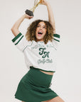 A woman wearing a white rugby-inspired polo shirt with green details and text that reads 'FORE ALL Golf Club'.