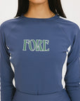 A female model wearing a long sleeve activewear top in blue and white with the text 'FORE' on the front, paired with a matching skirt.