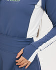 A female model wearing a long sleeve activewear top in blue and white with the text 'FORE' on the front, paired with a matching skirt and a visor with the text 'FORE'.