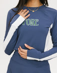 A female model wearing a long sleeve activewear top in blue and white with the text 'FORE' on the front, paired with a matching skirt and a visor with the text 'FORE'.