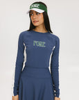 A female model wearing a long sleeve activewear top in blue and white with the text 'FORE' on the front, paired with a matching skirt and a visor with the text 'FORE'.