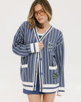 A woman wearing an oversized blue and white pinstripe cardigan with intarsia knit details and applique logos, paired with an indigo dress.