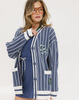 A woman wearing an oversized blue and white pinstripe cardigan with intarsia knit details and applique logos, paired with an indigo dress.