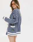 A woman wearing an oversized blue and white pinstripe cardigan with intarsia knit details and applique logos, paired with an indigo dress.