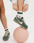A person standing on one leg placing a retro ankle sock into their shoe, with the sock displaying retro-inspired stripes in pink and green colors, and a logo 'FA' on the cuff.