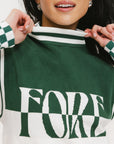 A woman wearing a sleeveless mock-neck sweater with a green and white intarsia knit design and the word 'FORE' written across the front.
