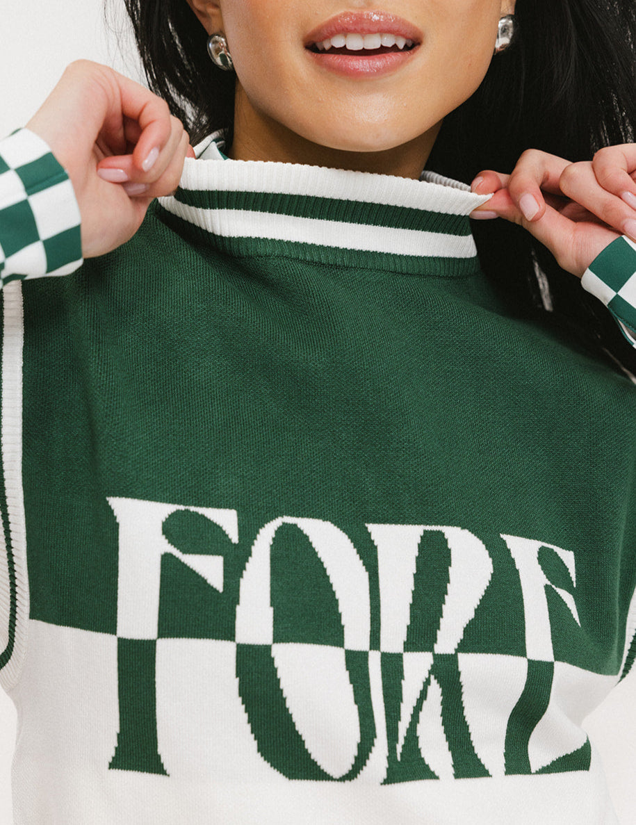 A woman wearing a sleeveless mock-neck sweater with a green and white intarsia knit design and the word 'FORE' written across the front.