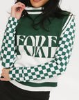 A woman wearing a sleeveless mock-neck sweater with a green and white intarsia knit design and the word 'FORE' written across the front.