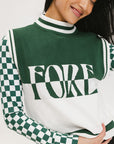 A woman wearing a sleeveless mock-neck sweater with a green and white intarsia knit design and the word 'FORE' written across the front.