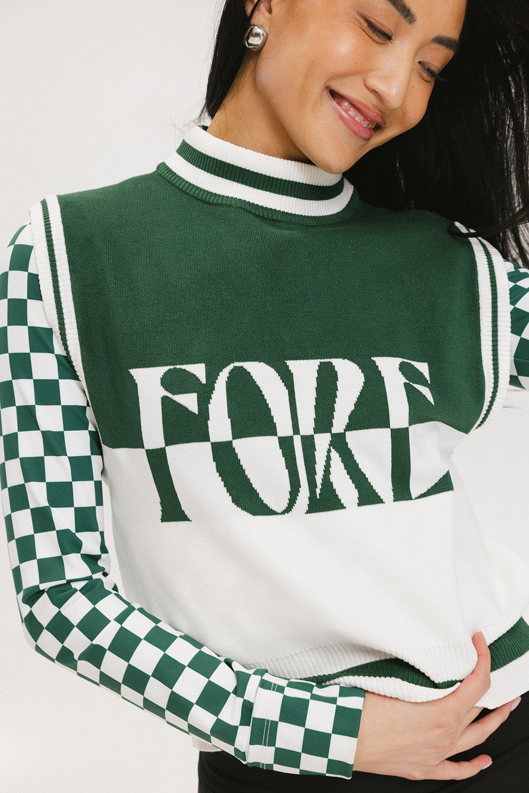 A woman wearing a sleeveless mock-neck sweater with a green and white intarsia knit design and the word 'FORE' written across the front.