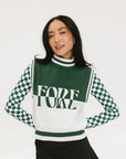 A woman wearing a sleeveless mock-neck sweater with a green and white intarsia knit design and the word 'FORE' written across the front.
