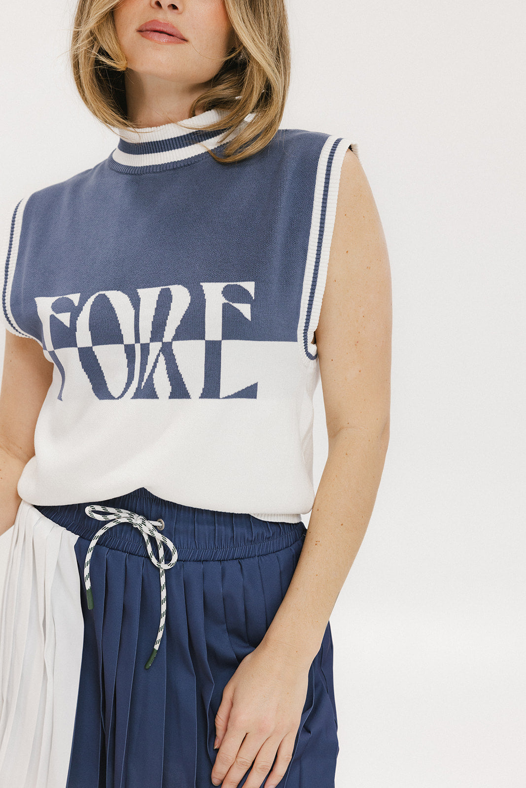 A model wearing an indigo and white intarsia mock neck, sleeveless Fore Sweater.