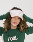 A woman wearing a pink and white 'FORE' visor and a green long-sleeved shirt.