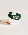 A hand holding a white and green two-toned visor with the word 'FORE' printed on the front.