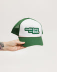 A hand holding a trucker hat with 'Double Bogeys Are Hot' text embroidered on the front, featuring a white mesh back and green visor.