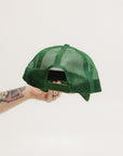 A hand holding a white and green trucker hat with the brim facing up.