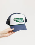 A hand holding a trucker hat with 'Double Bogeys Are Hot' text embroidered on the front, featuring a white panel with green text and a black mesh back.