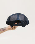 A hand holding a trucker hat with a white front and indigo blue mesh back.